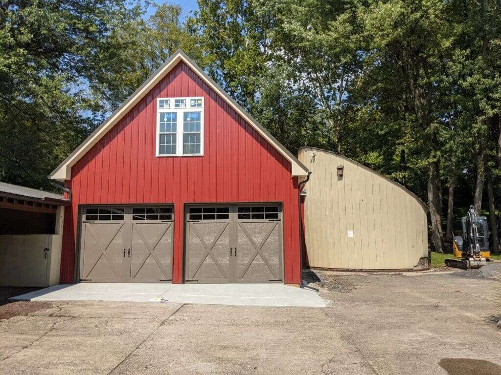 Two-story garage with red board-and-batten siding, carriage doors, upper loft windows, and attached lean-to