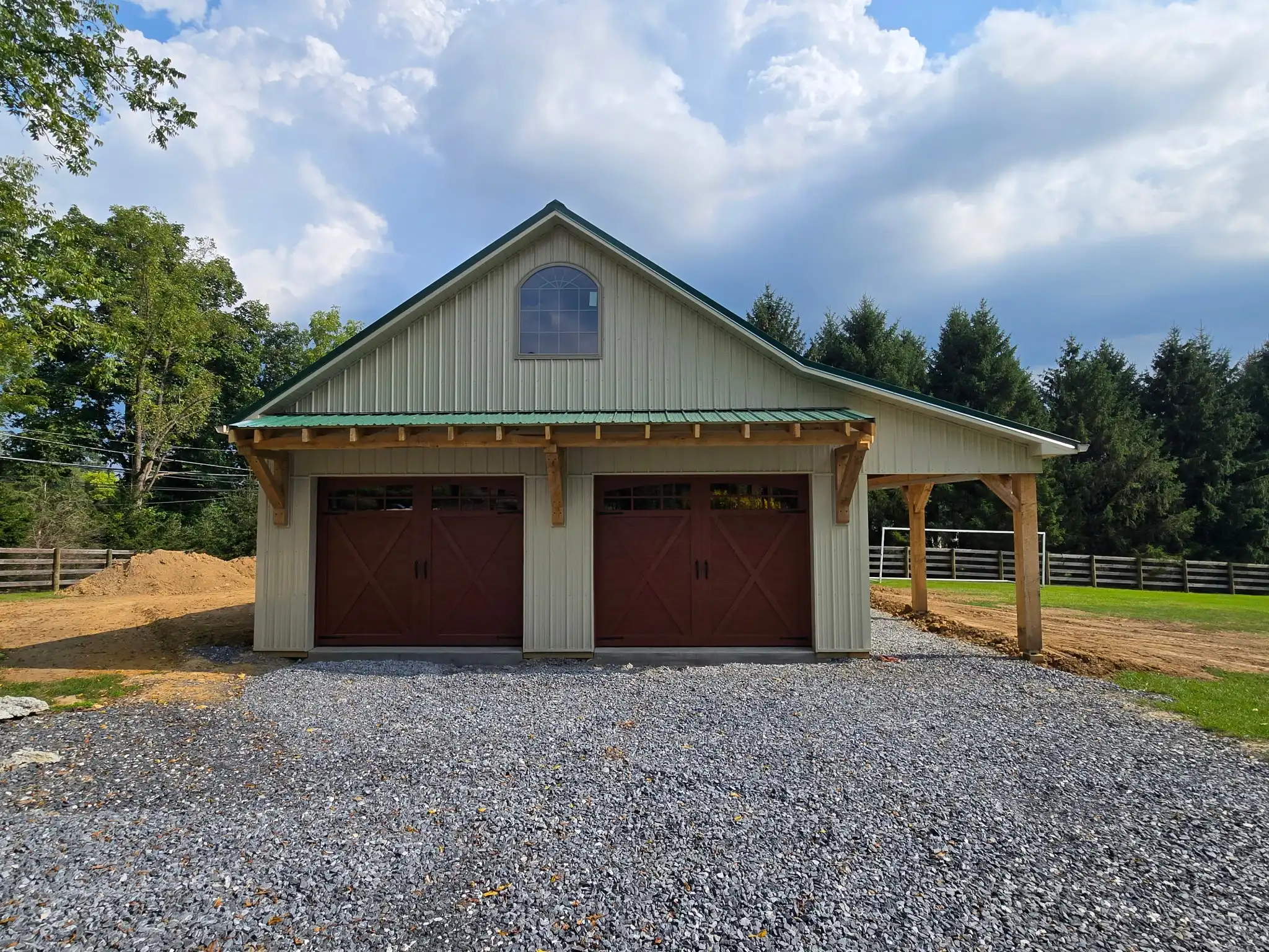 Two-story garage with sage green metal siding, green metal roof, timber-framed overhang above two brown carriage-style doors, arched loft window, and open lean-to bay
