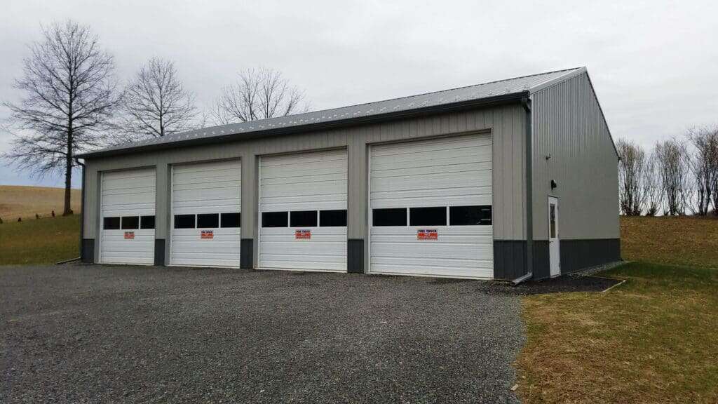 Grey post-frame fire truck storage building with four large overhead doors labeled Fire Truck