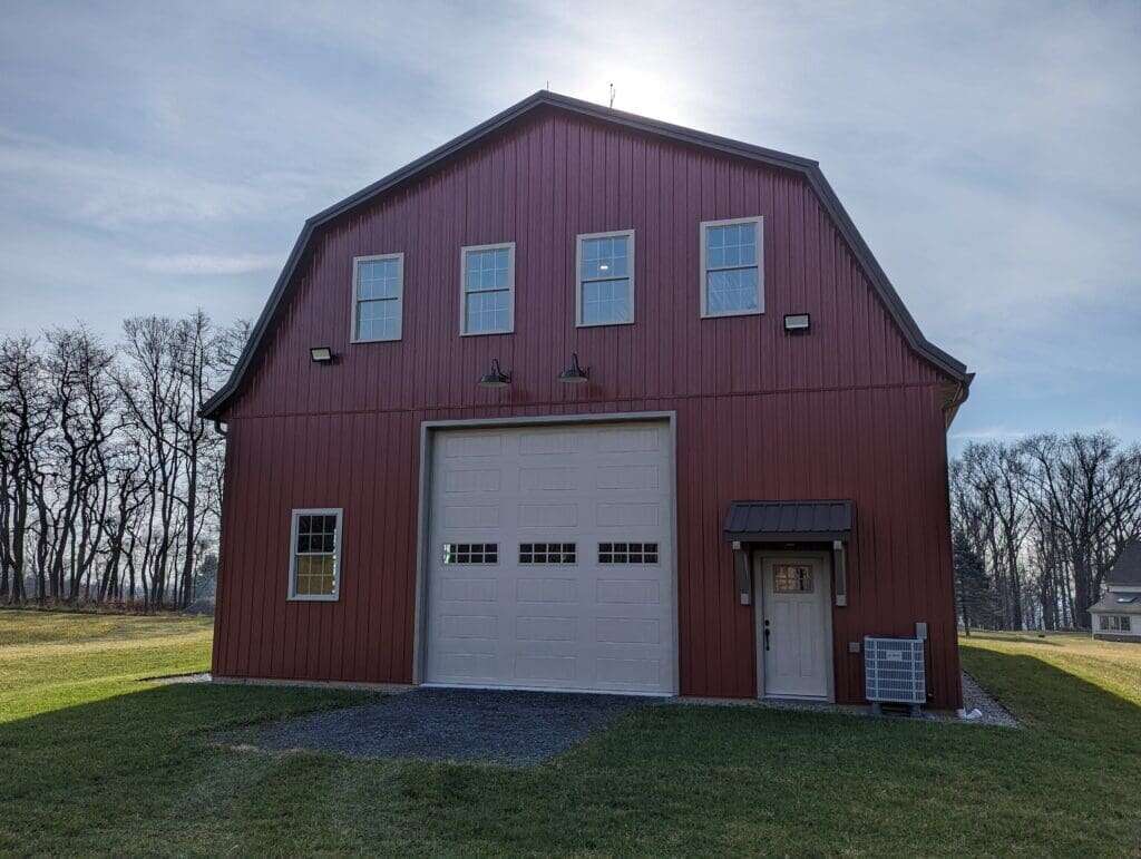 Two-story gambrel-style garage with red metal siding, white overhead door, and upper loft windows with barn lighting