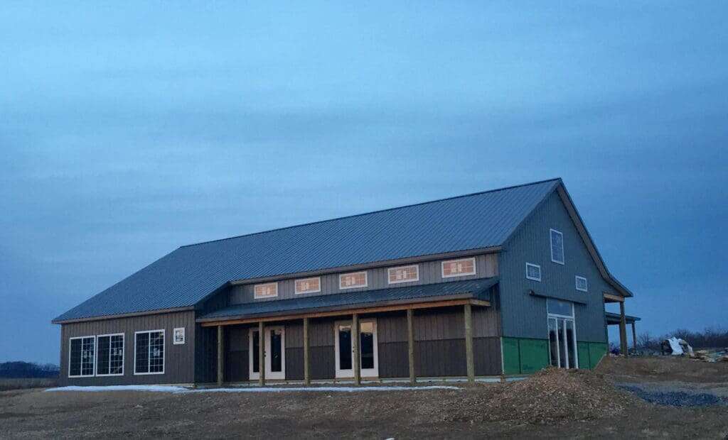 Large barndominium under construction with grey-brown metal siding, standing seam roof, and full-length covered porch with timber posts