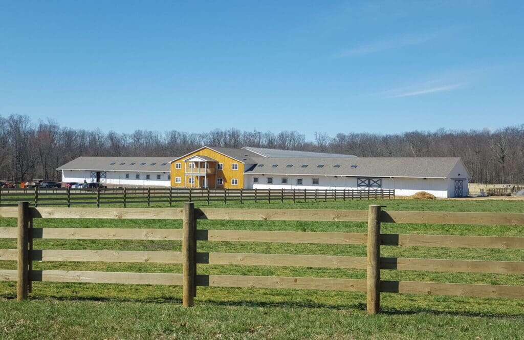 Completed equestrian facility from pasture showing full arena, yellow barn house, and fenced paddocks