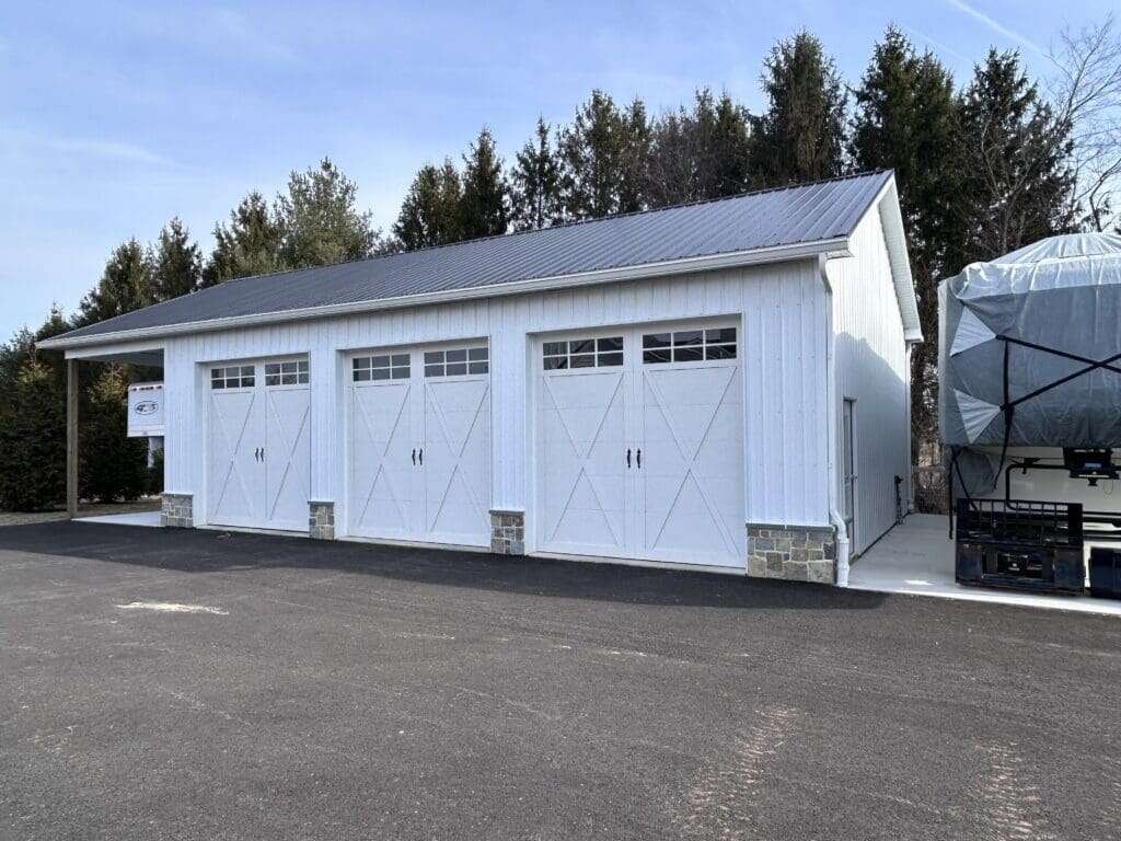 White three-bay post-frame garage with charcoal metal roof, carriage-style overhead doors, and stone veneer corner accents