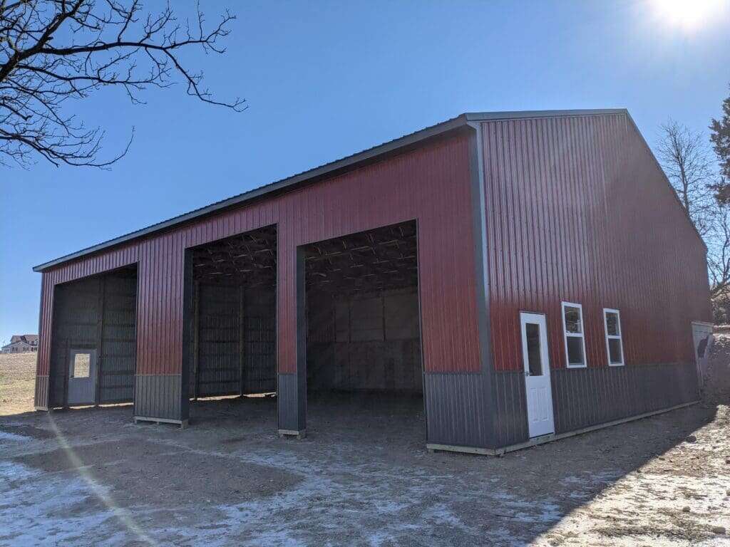 Large three-bay post-frame garage with red and charcoal two-tone metal siding and three open overhead bays