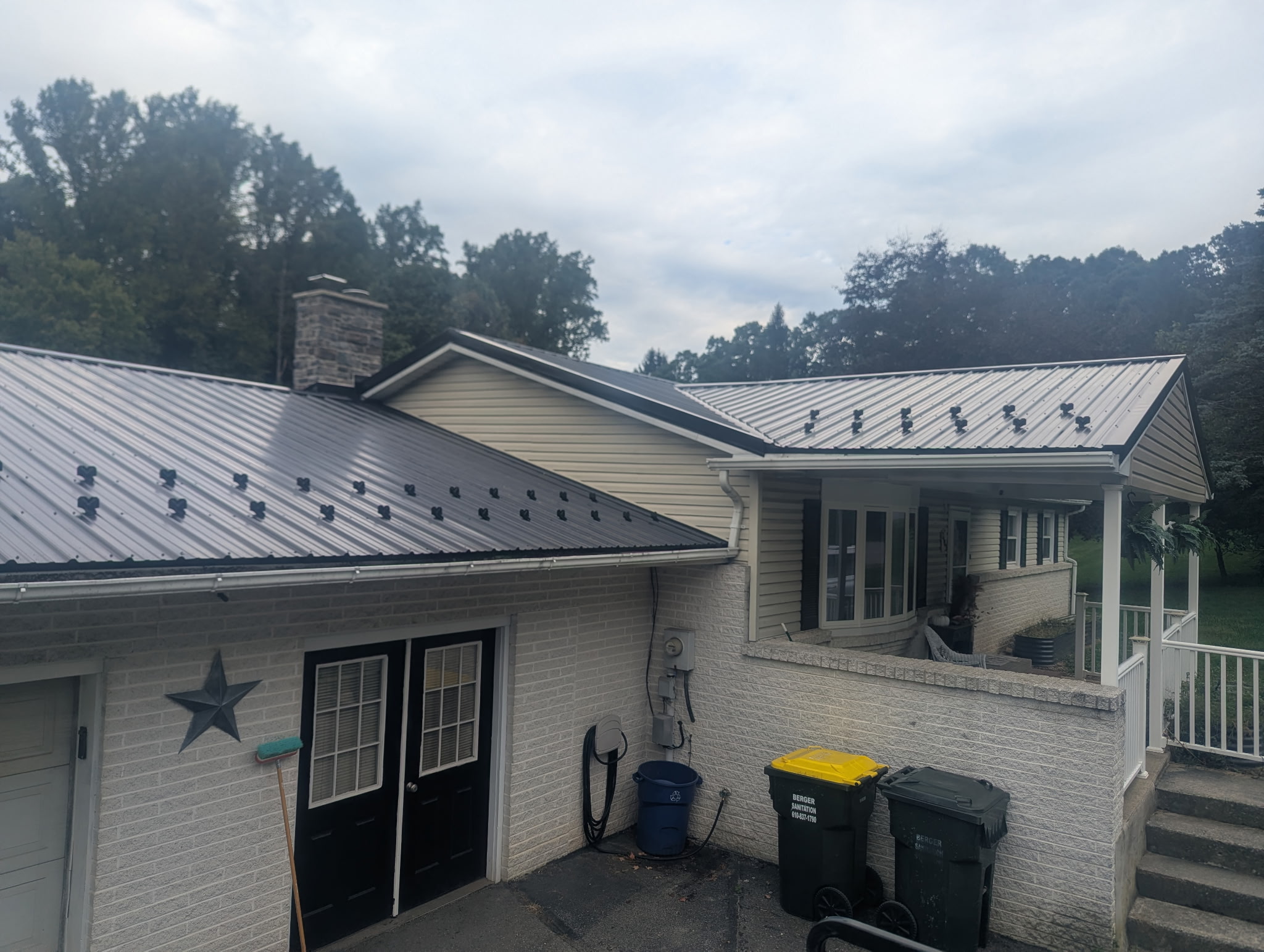 Ranch-style home with new dark gray standing-seam metal roof, stone chimney, tan vinyl siding, and covered front porch with white columns