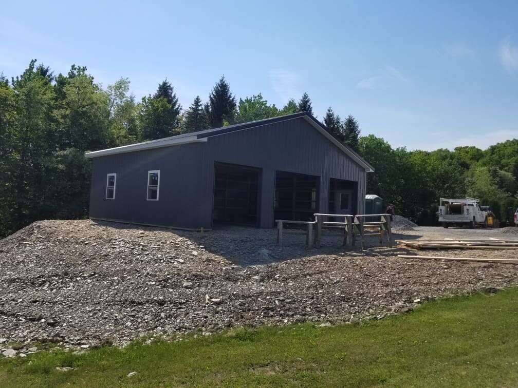 Dark grey post-frame garage with two large overhead bays under construction on a wooded lot