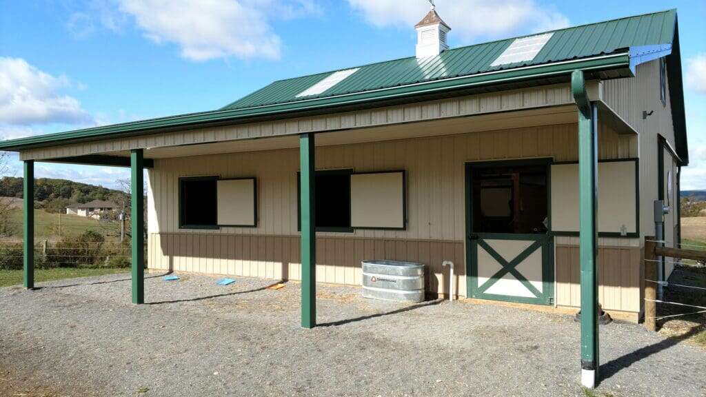 Front elevation of tan horse barn with green posts, covered overhang, and stall windows