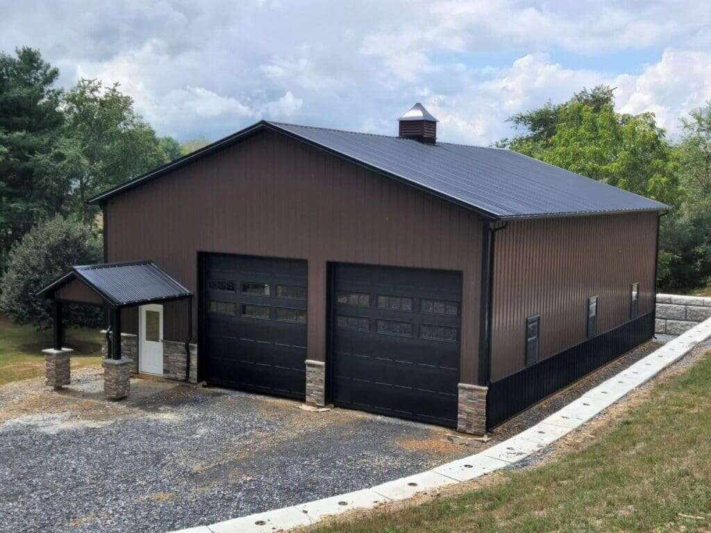 Post-frame garage with dark brown metal siding, black metal roof, stone veneer wainscot, and decorative stone entry pillars
