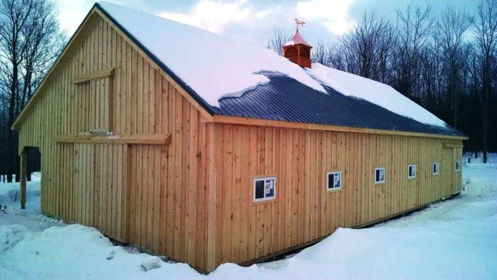 Completed post-frame storage barn with natural cedar siding, dark metal roof, cupola with weathervane, and small windows in winter snow