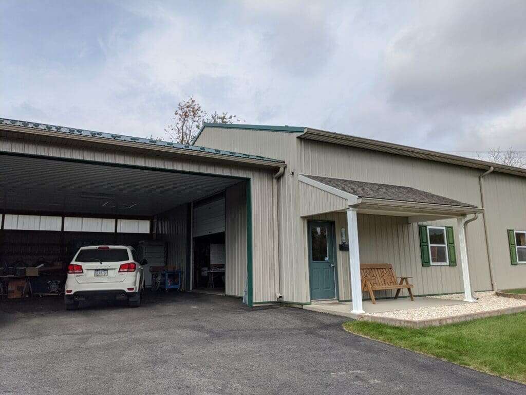 Commercial post-frame building with tan siding, green metal roof, open garage bay, and office entry with teal door