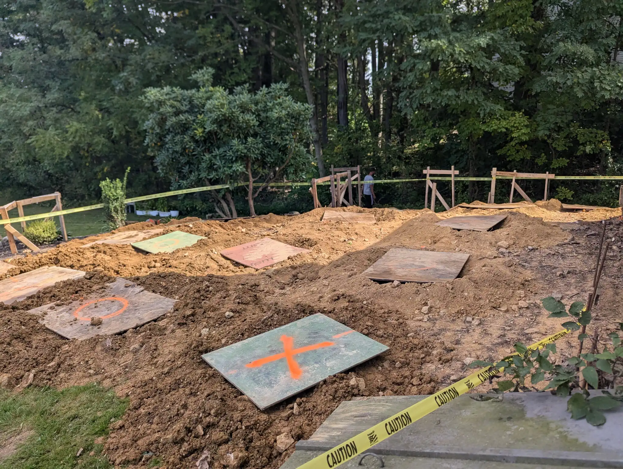 Excavated backyard construction site with plywood footer covers, orange marking paint, and caution tape staked around the perimeter