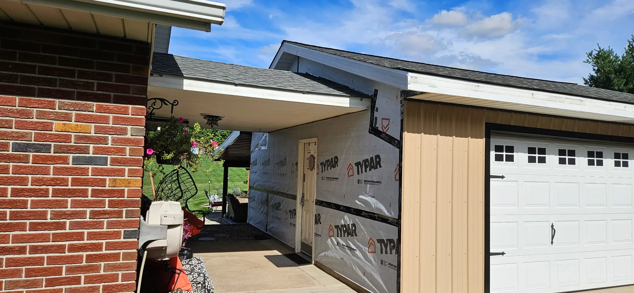 Brick ranch home mid-residing with white Typar house wrap on the walls, tan metal siding installed on the attached garage, and a white carriage-style garage door